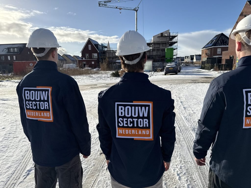 Drie vakmensen van Bouwsector Nederland in blauwe jassen bij een bouwplaats met hijskraan